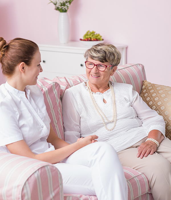 Caregiver and elderly woman smiling and talking while seated together on a couch at home.